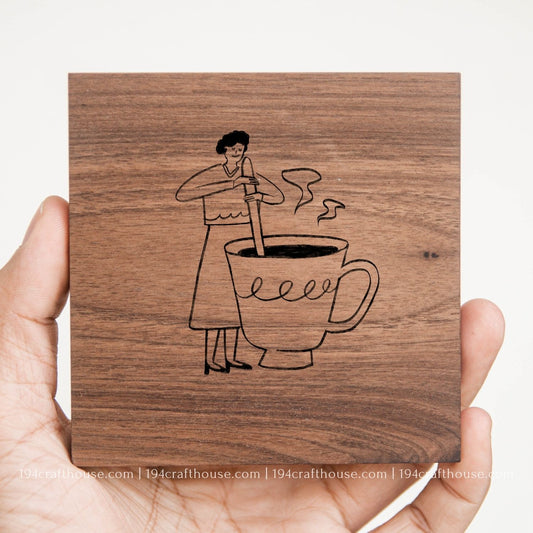 Wooden coaster with a engraved illustration of a person making coffee held by a hand.
