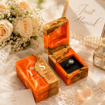 Personalized orange resin and wood ring box engraved with initials and date set in a wedding scene with flowers and candles.