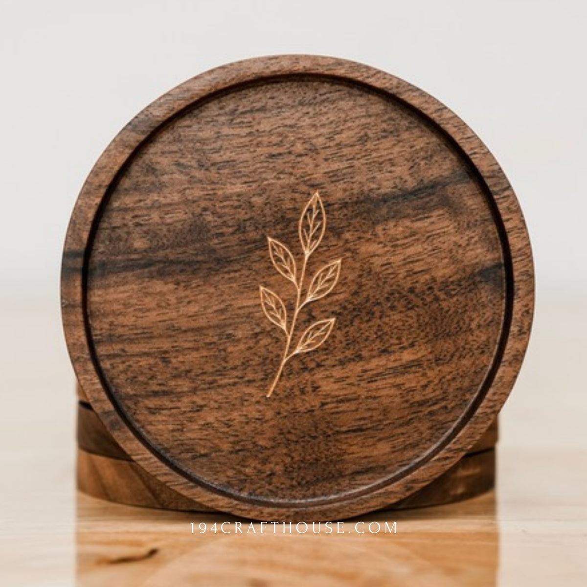 Front view of a round walnut coaster engraved with a small leaf sprig, standing on a surface