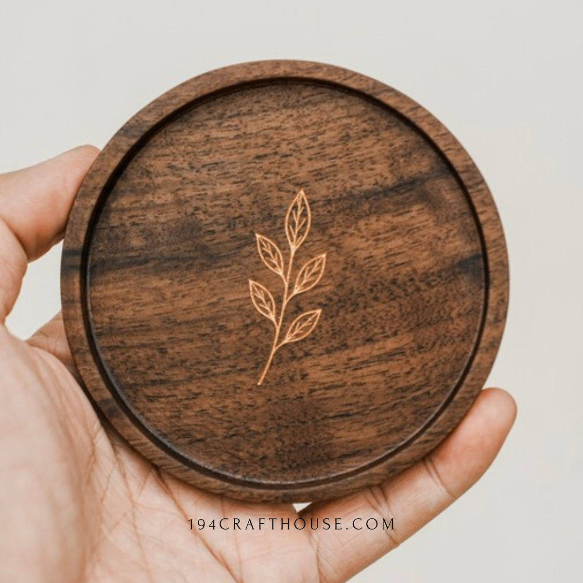 Round walnut coaster with engraved leaf sprig held in a hand against a light background