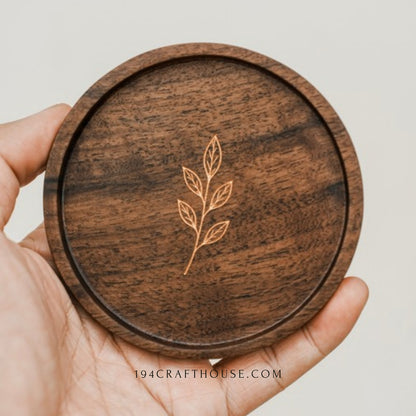 Round walnut coaster with engraved leaf sprig held in a hand against a light background