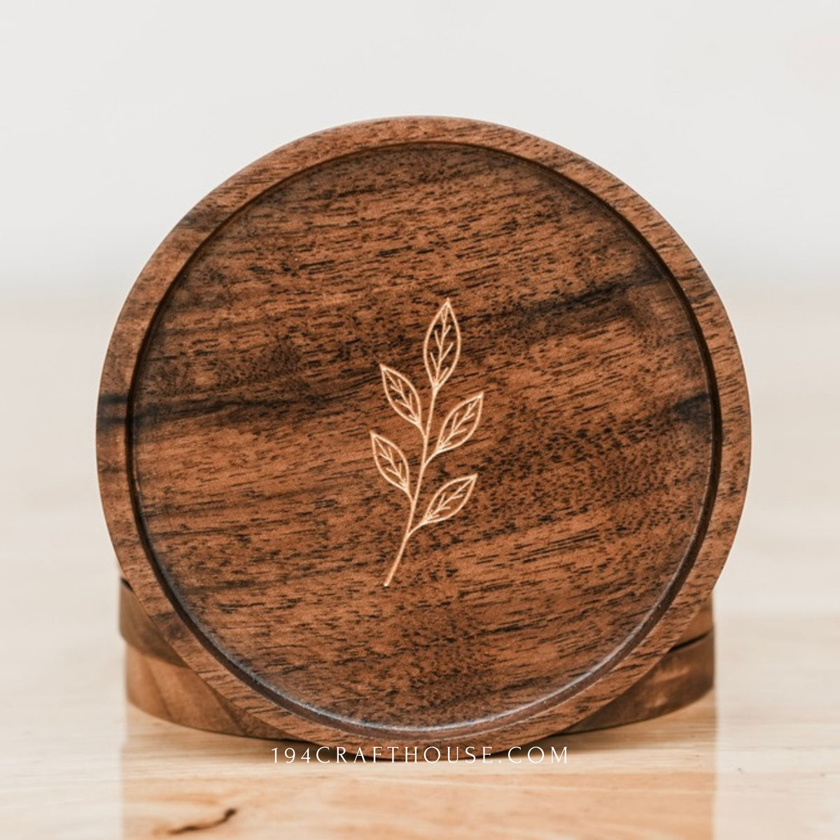 Round walnut coaster with leaf sprig engraving displayed upright with another coaster behind it