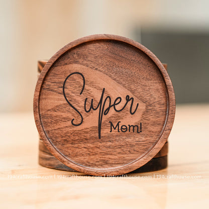 A round wooden coaster with the words 'Super Mom!' engraved in black script, displayed on a light wooden surface with a soft-focus background.