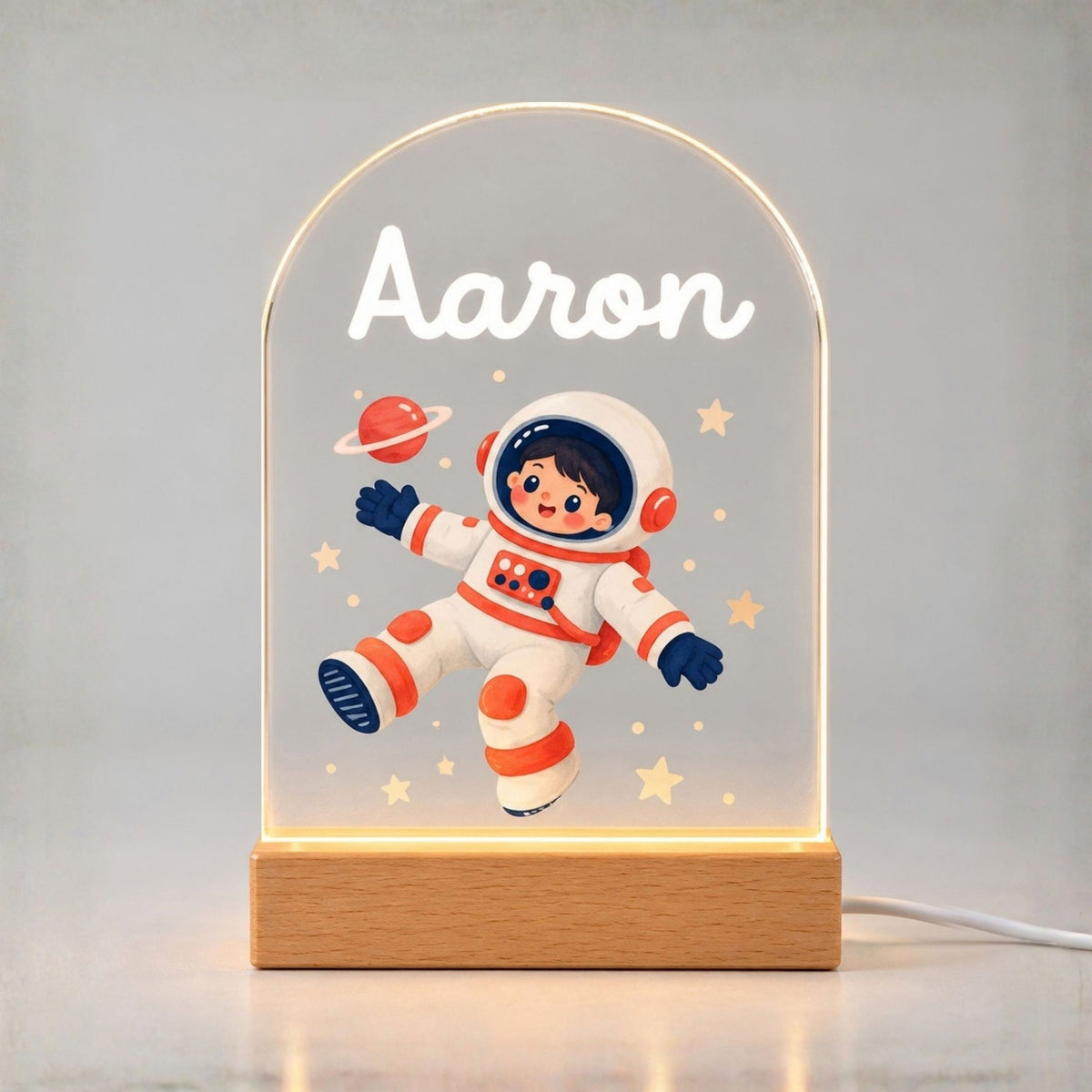 Glowing arched acrylic night light shows Aaron, a cartoon astronaut, stars, and a ringed planet above a wooden base.