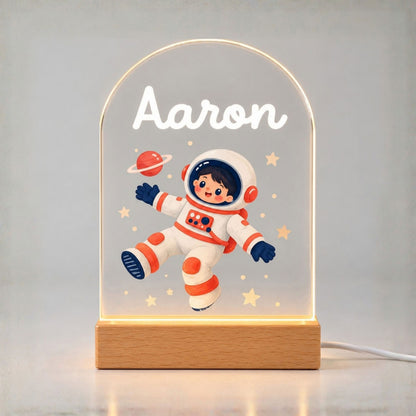 Glowing arched acrylic night light shows Aaron, a cartoon astronaut, stars, and a ringed planet above a wooden base.