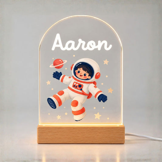 Glowing arched acrylic night light shows Aaron, a cartoon astronaut, stars, and a ringed planet above a wooden base.