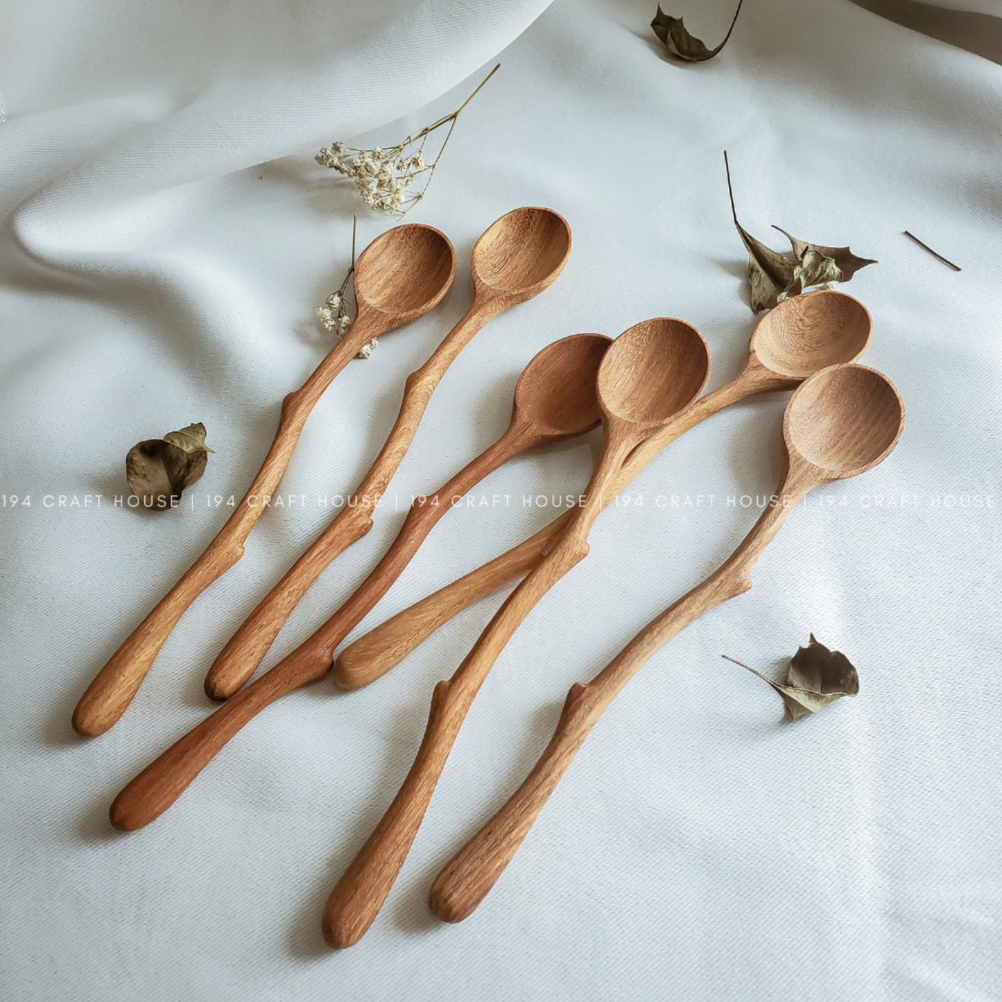 A set of eight wooden spoons with a natural olive oil finish, displayed on a white fabric surface with some dried leaves and petals scattered around.