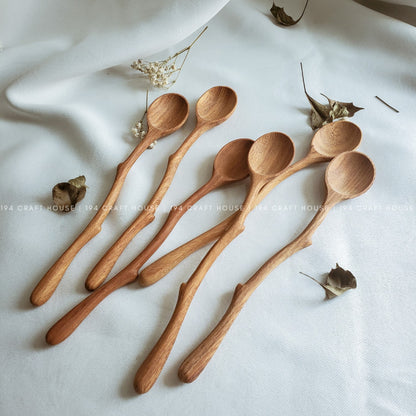 A set of eight wooden spoons with a natural olive oil finish, displayed on a white fabric surface with some dried leaves and petals scattered around.