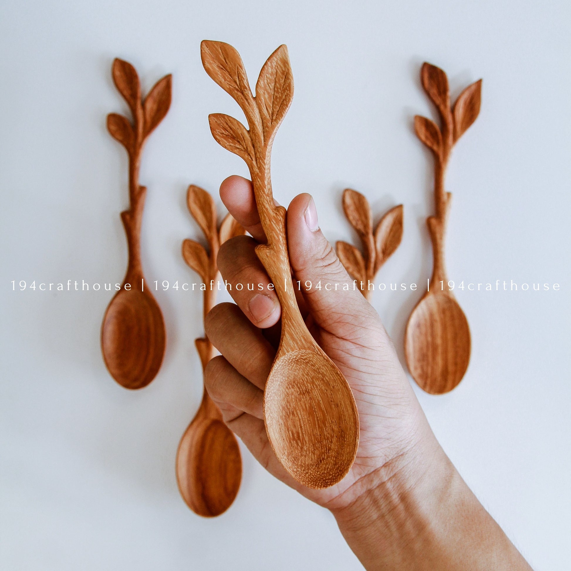 A hand holds a carved wooden spoon with a leafy branch handle, while four similar wooden spoons are displayed upright in the background against a plain light wall.