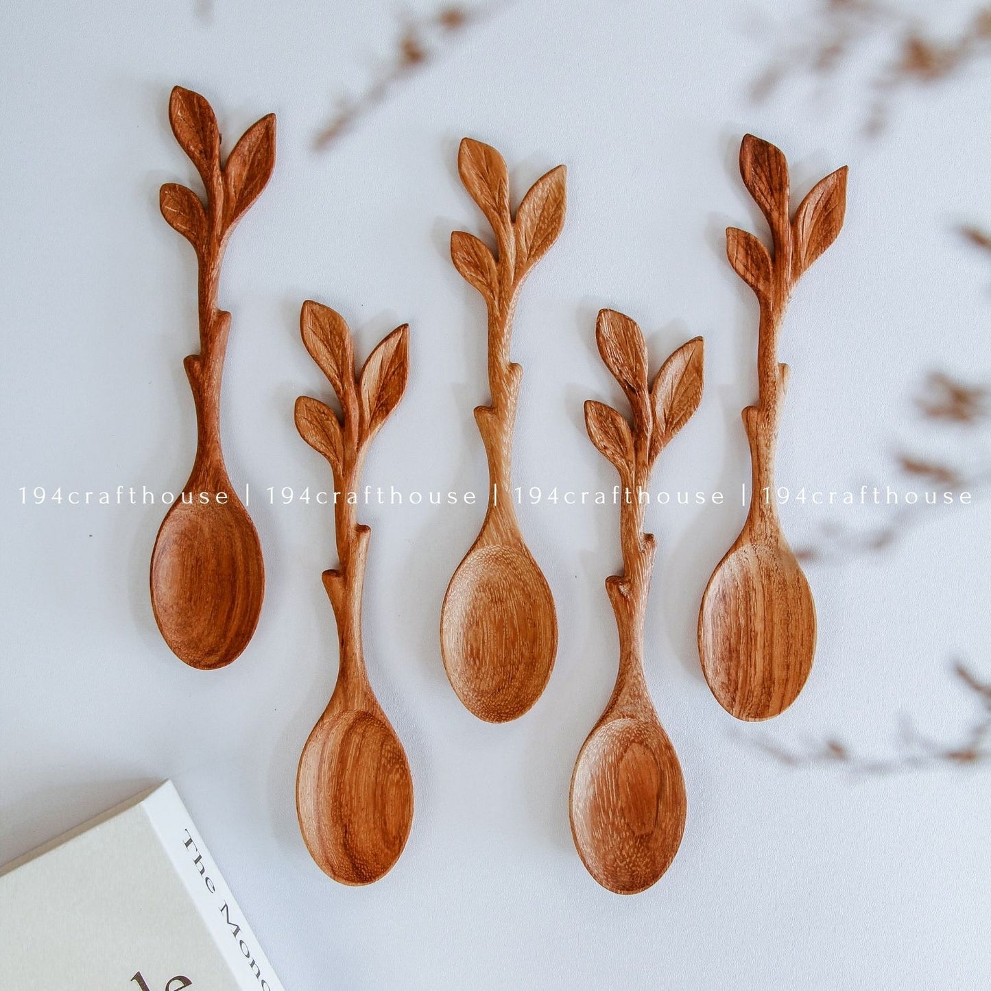 Five wooden spoons with leaf-shaped handles, arranged on a light background with a book corner visible in the lower left.