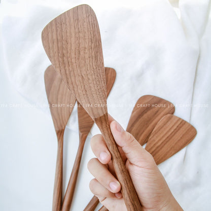 A walnut wood cooking spatula on hand with the background set of handcrafted walnut wood spatulas with unique natural grain