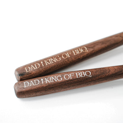 Angled close-up of two walnut handles bearing “DAD I KING OF BBQ” engravings