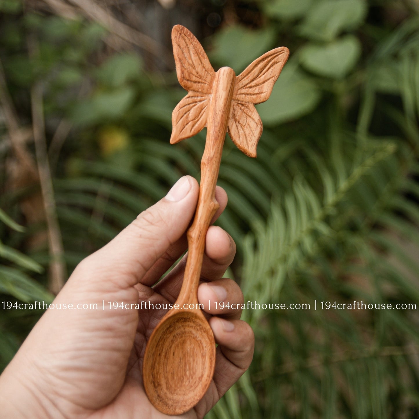 Hand-carved Butterfly wooden spoon crafted from doussie wood, featuring a sculpted butterfly handle hold by hand on a grenn tree background.