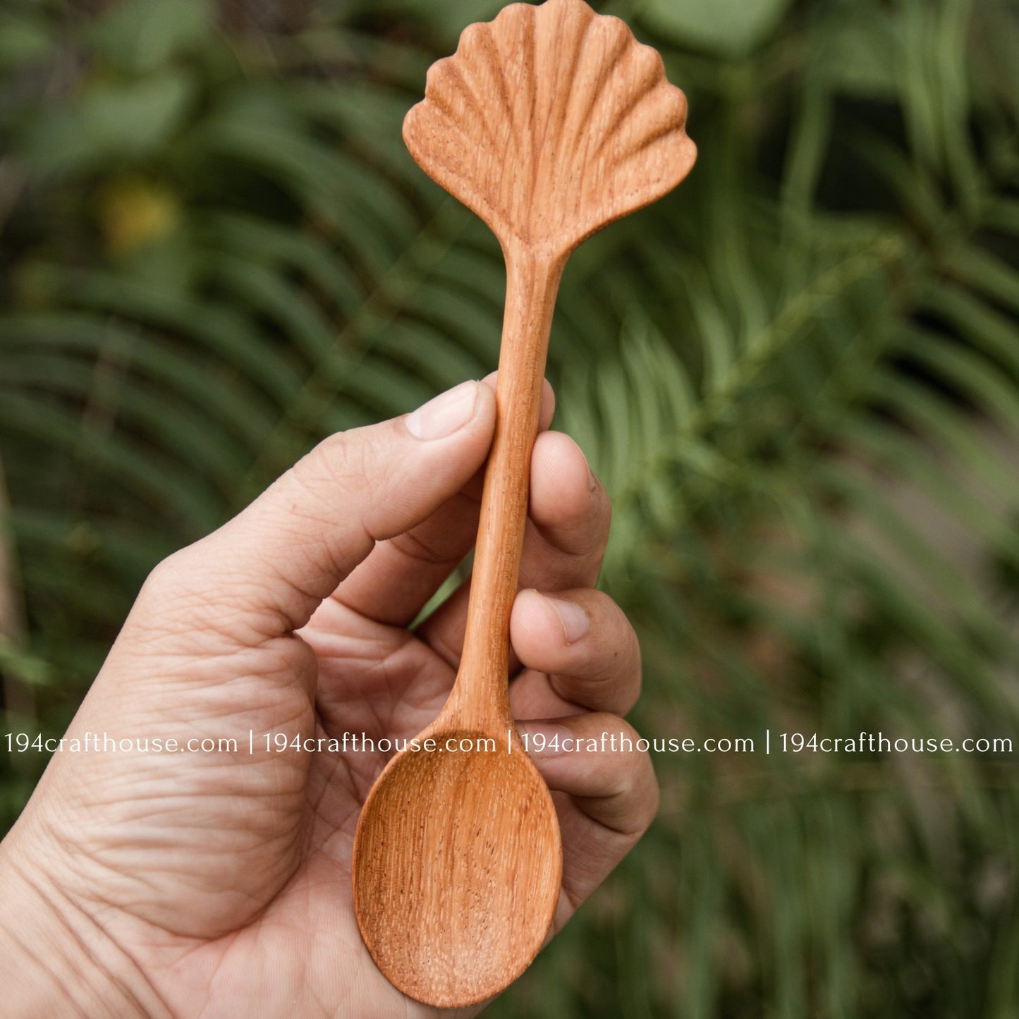 Hand-carved seashell wooden spoon crafted from doussie wood, featuring a sculpted mushroom cap handle hold by hand on a green tree background.