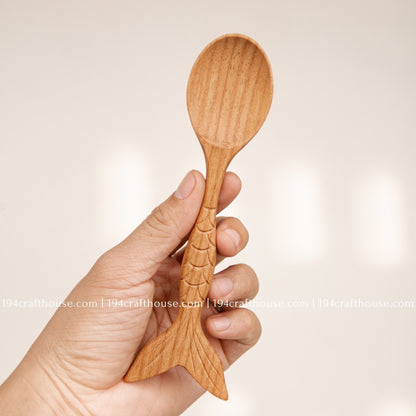 Hand holding a Doussie wood spoon to show its oval bowl, carved scale details and fish-tail handle.