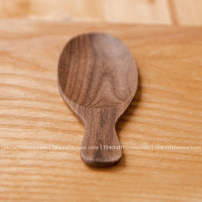 Single short-handle walnut wood tea scoop on a natural wood surface, showing its deep oval bowl.
