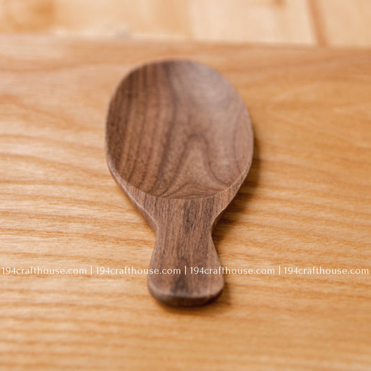 Single short-handle walnut wood tea scoop on a natural wood surface, showing its deep oval bowl.