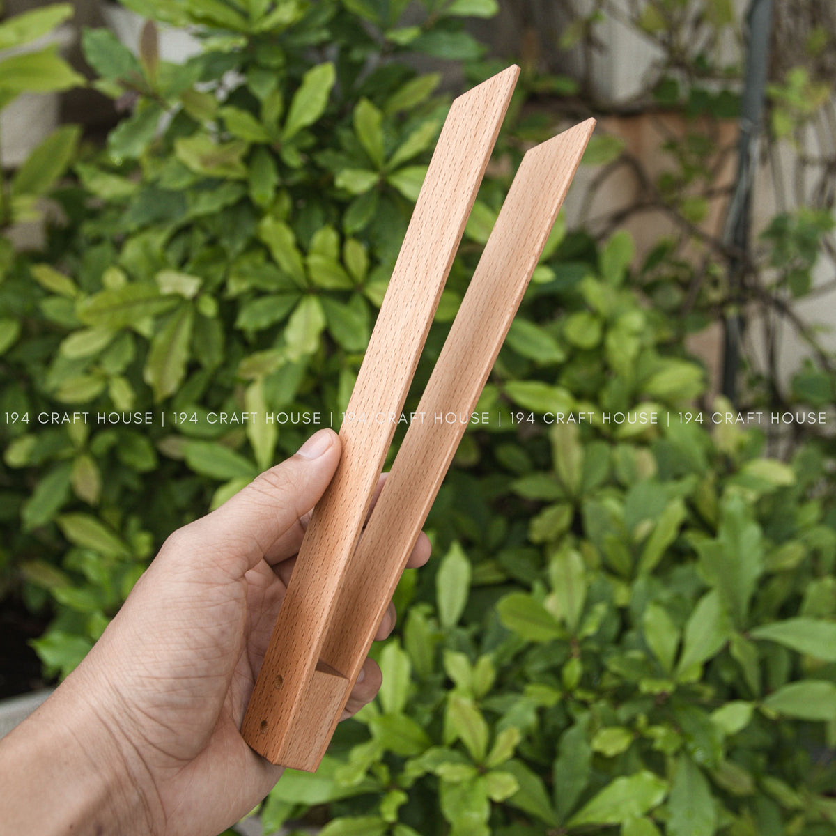 A hand holds the open wooden salad tongs in front of leafy green plants.