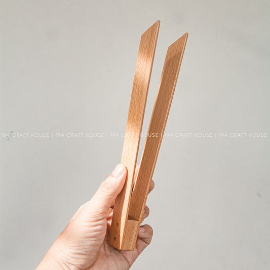 A hand holds the open beech wood salad tongs upright against a plain background.