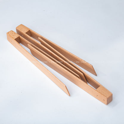 The open wooden salad tongs lie diagonally on a light surface, showing both long arms.