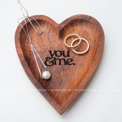 Heart-shaped wooden tray with 'you & me' engraving, necklace, and rings on a white background.