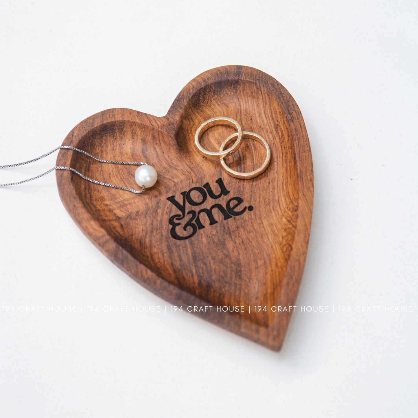 Wooden heart-shaped tray with 'you & me' engraving, holding a necklace and two rings on a white background.