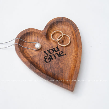 Wooden heart-shaped tray with 'you & me' engraving, holding a necklace and two rings on a white background.