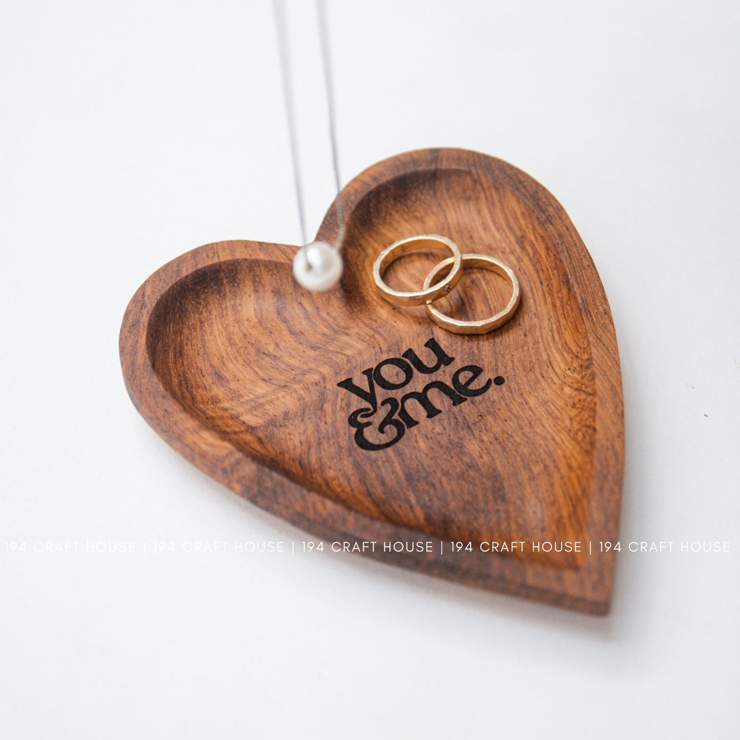Wooden heart-shaped dish with 'you & me' engraving and two gold rings on a white background.