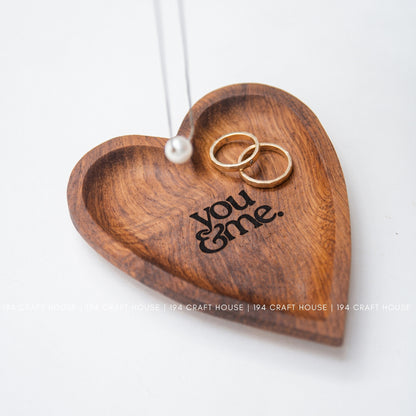 Wooden heart-shaped dish with 'you & me' engraving and two gold rings on a white background.