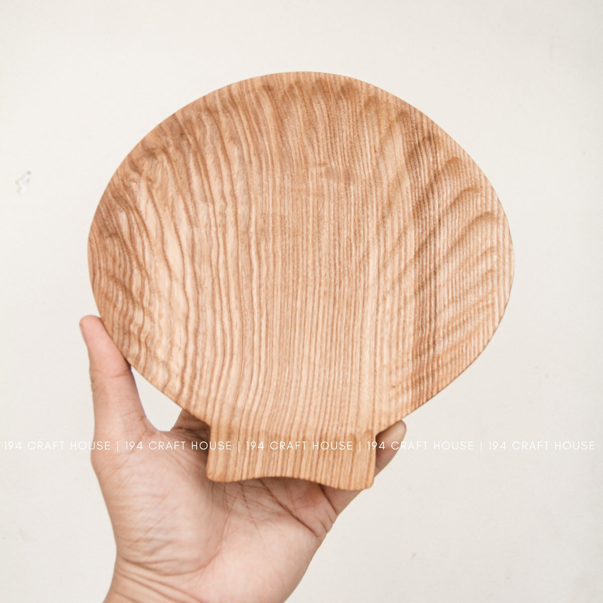 A hand holds the shell-inspired ash wood tray upright, showing its rounded shape and projecting tab.