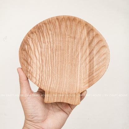 A hand holds the shell-inspired ash wood tray upright, showing its rounded shape and projecting tab.
