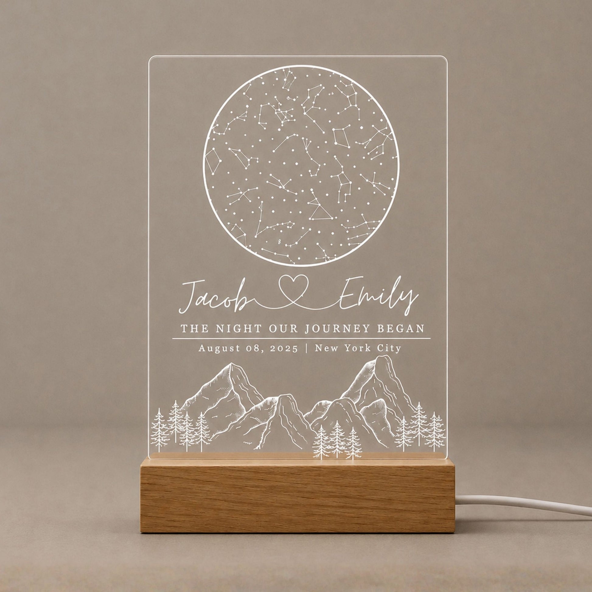 Studio front view of unlit clear acrylic circular star map plaque engraved with couple names and mountain scenery mounted on a natural beech wood base.
