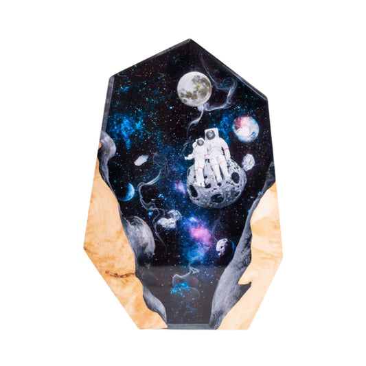 A studio photograph of a hexagonal resin lamp against a plain white background. The lamp contains a miniature scene of two astronauts sitting together on a cratered moon, surrounded by a swirling blue and pink galaxy with distant planets