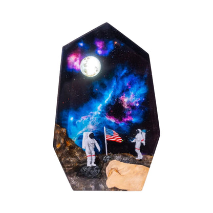 A high-resolution photo of a hexagonal resin lamp against a plain white background. The design depicts a moon landing scene with two astronauts and an American flag on a rocky lunar surface