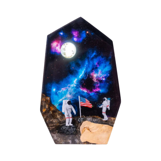 A high-resolution photo of a hexagonal resin lamp against a plain white background. The design depicts a moon landing scene with two astronauts and an American flag on a rocky lunar surface