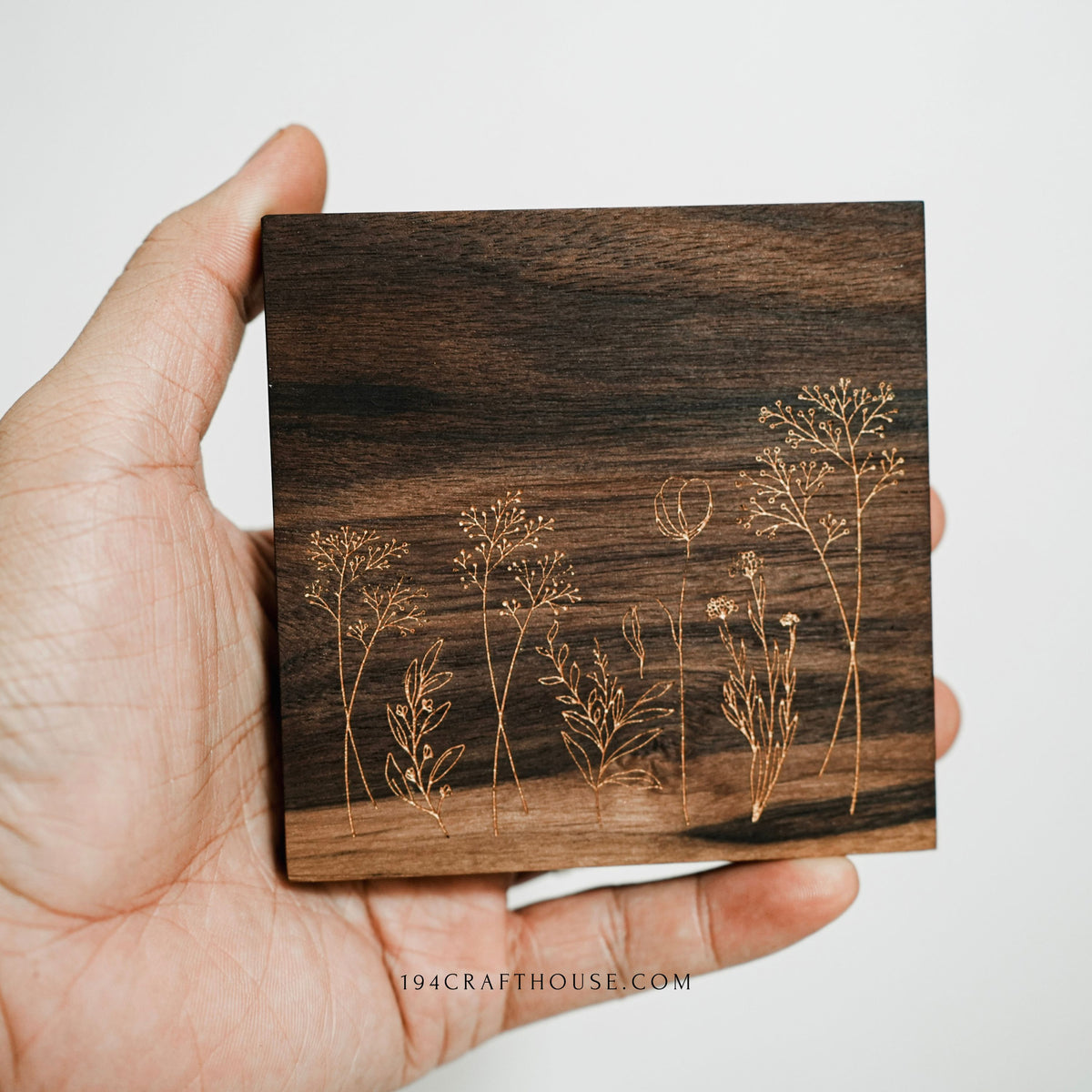 A hand holding a square walnut wood coaster with an engraved wildflower botanical design.