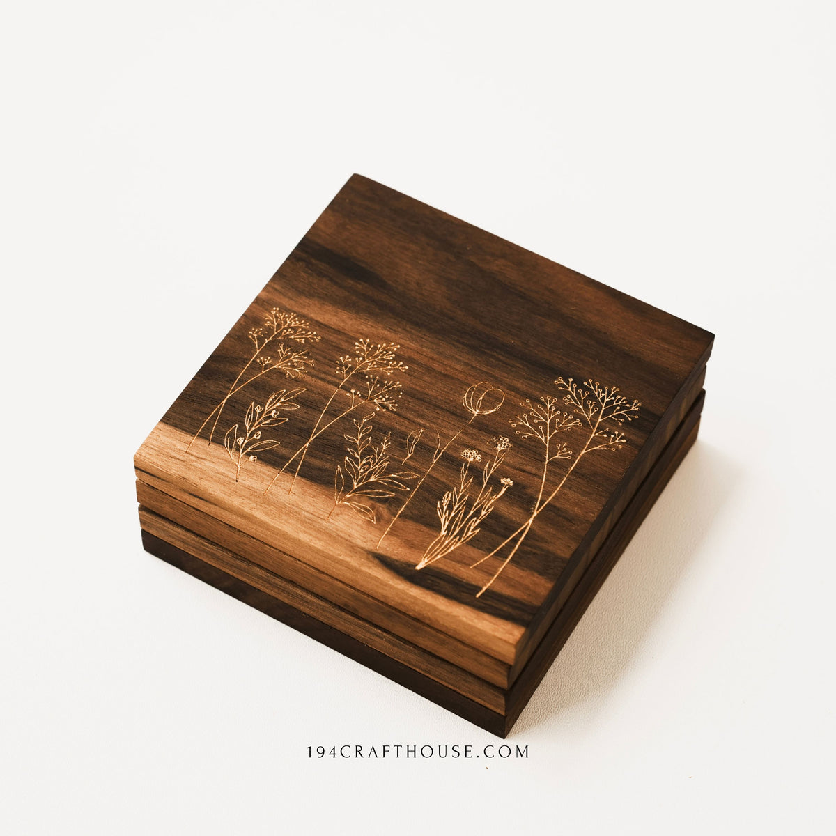 Stack of walnut wood coasters with wildflower engravings viewed from the front on a white surface.