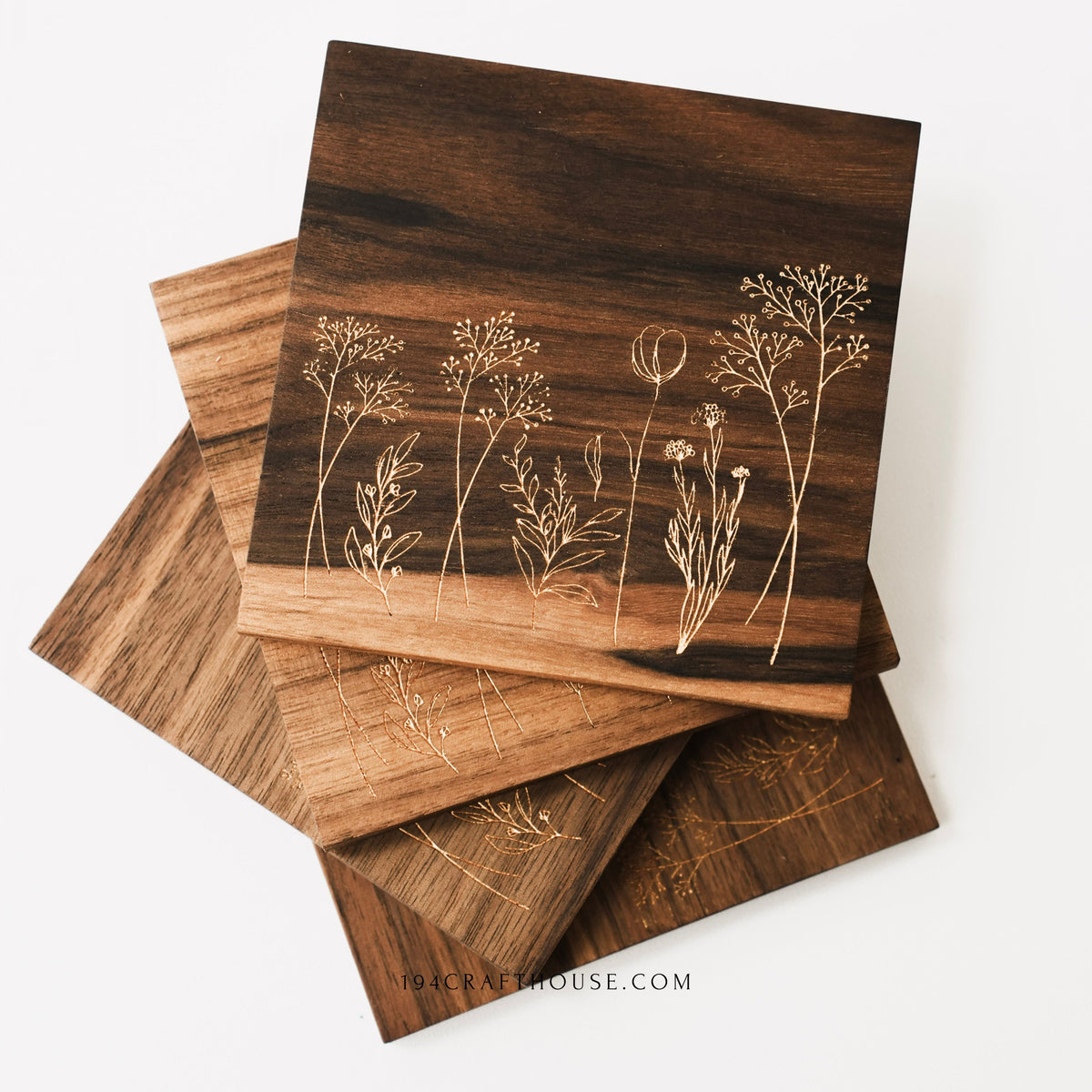 Four walnut wood coasters with engraved wildflower designs stacked in a fan shape on a white background.